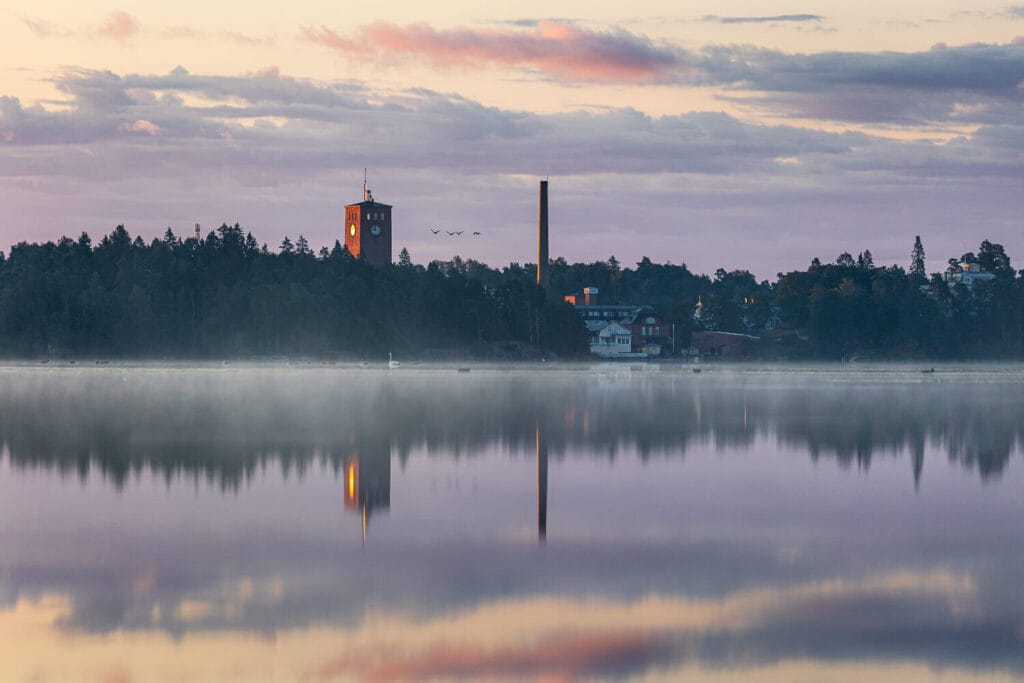 Sumuinen auringonnousu Littoistenjärvellä, Verkarannan tehdasrakennukset ja kellotorni heijastuvat tyynestä vedestä.