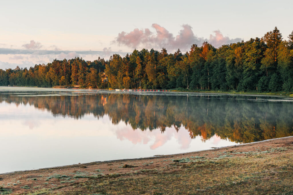 Littoistenjärven hiekkaranta ja tyyni järvimaisema aamunvalossa, metsä heijastuu veden pinnasta.